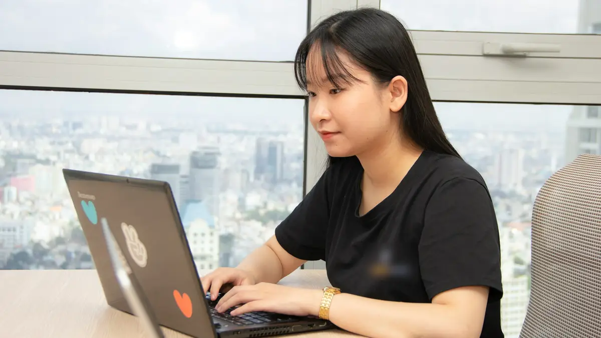 Cô gái trẻ ngồi gõ máy tính xách tay bên cửa kính văn phòng trên cao, phía sau là nhà cửa thành phố